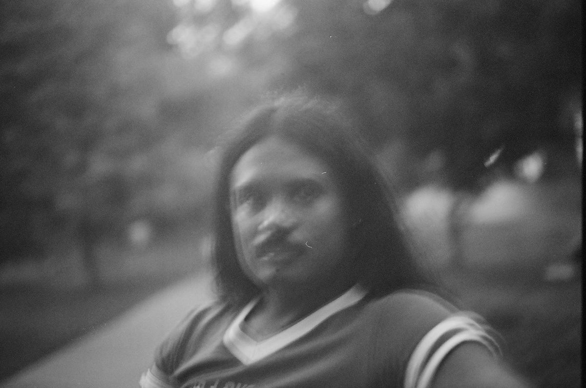 A black-and-white portrait of a person with long dark hair and a pale-collared shirt, looking to camera, background out of focus.